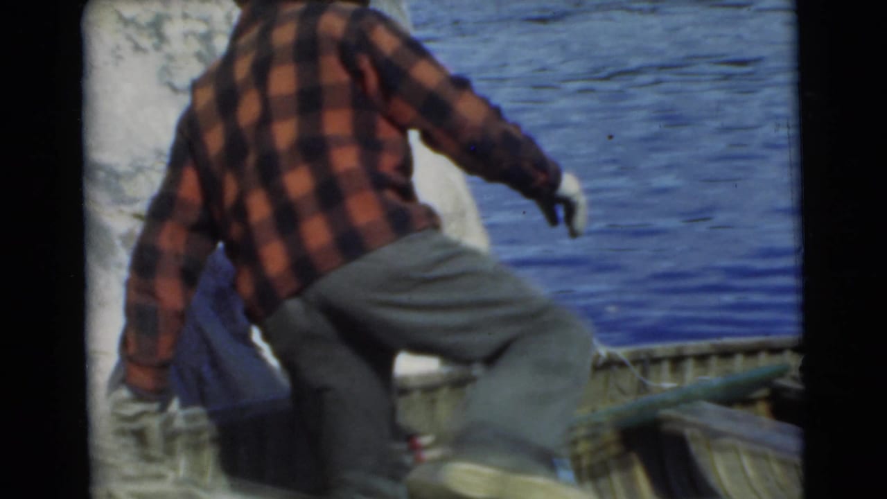 In 1956 Miami home-movie footage, a man in a red plaid shirt steps into or beside a wooden boat along blue lake water.