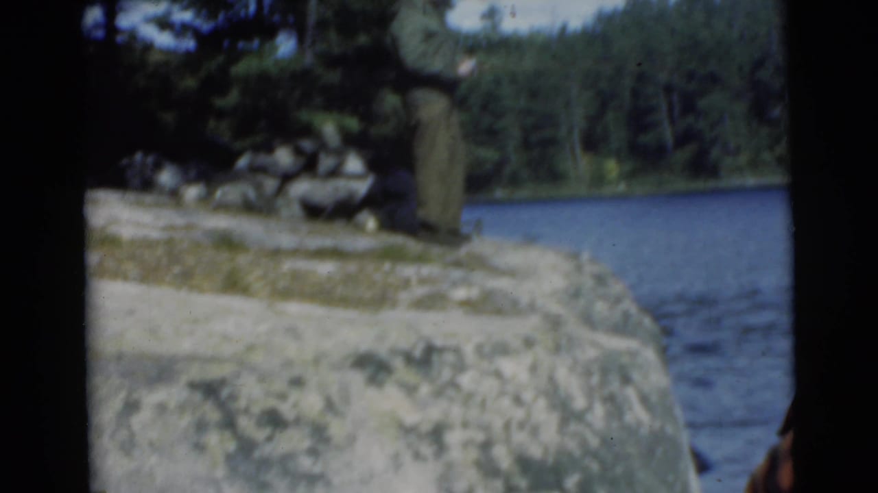 Man stands on a rocky lakeshore with water and trees behind him.