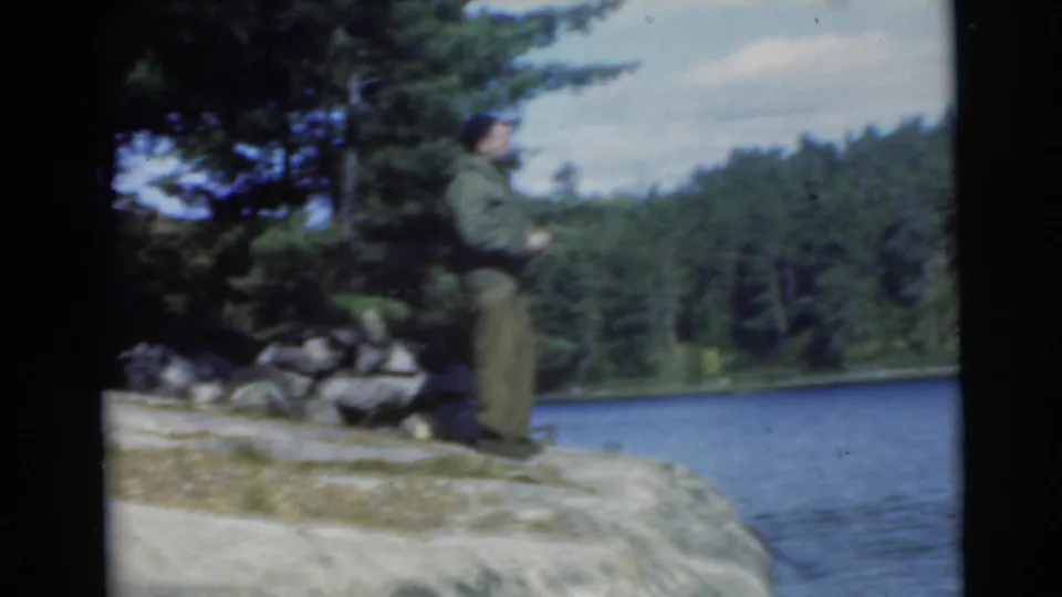 A man appears to fish from a rocky lakeshore in the 1956 Miami home-movie clip, with a wooded bank in the background.