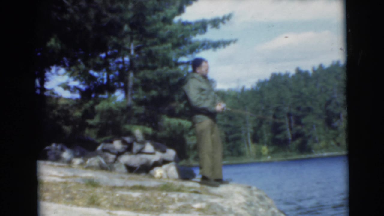 Man standing on a rocky lakeshore beside trees and blue water.