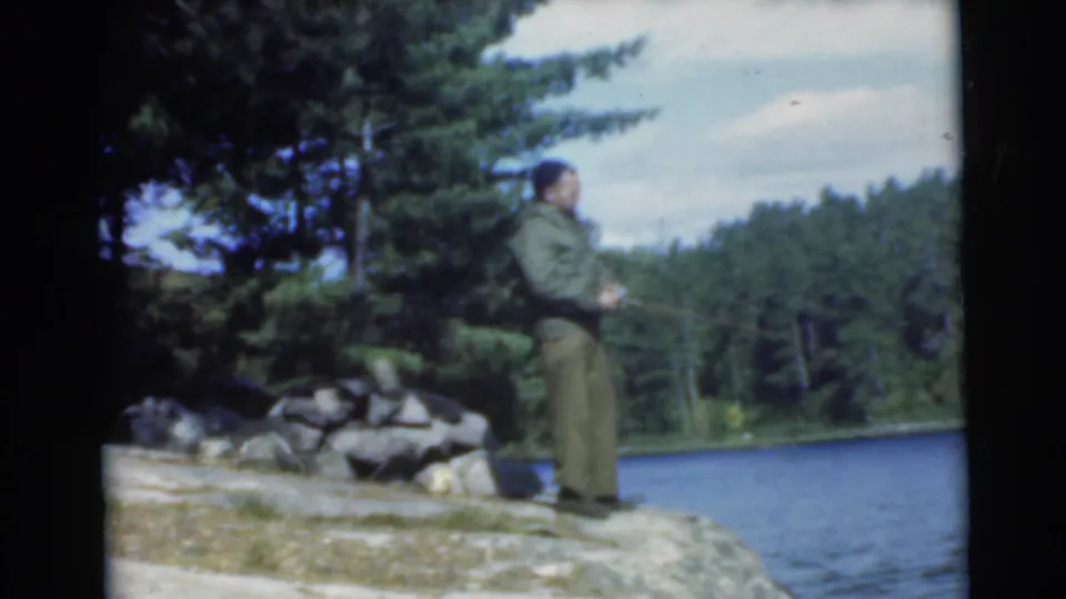 Adult man in a green jacket stands on a rocky lakeshore near pine trees and blue water in 1956 home-movie footage identified as Miami, Florida.