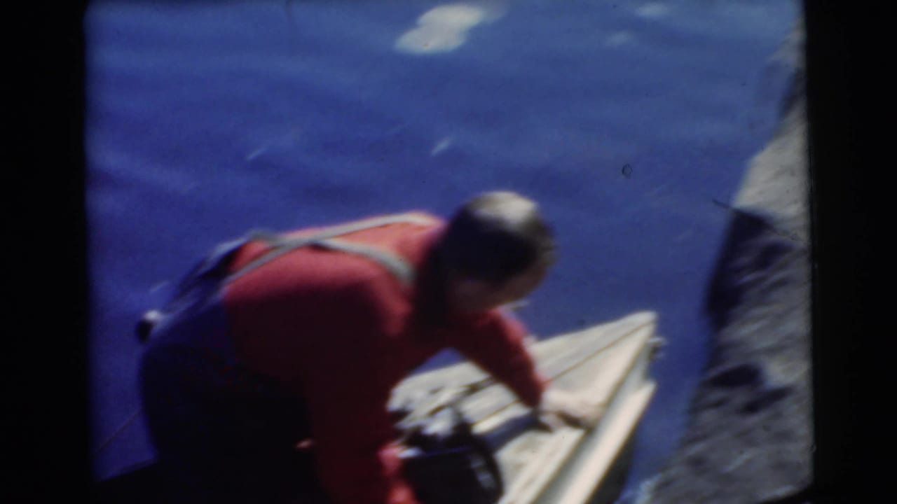 Man in a red shirt leans over a wooden canoe on blue water.