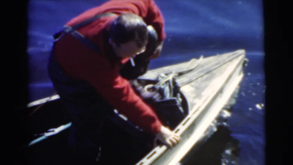 Adult man in red bends over the side of a wooden canoe, adjusting items as the boat sits beside deep blue water.