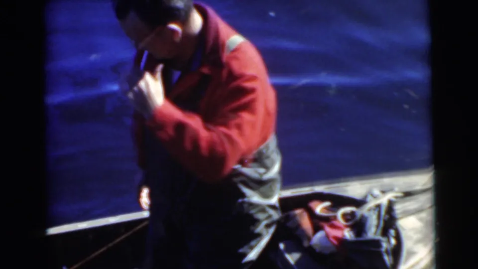 Adult man in a red shirt stands in or beside a canoe with bags and rope-like gear visible against the lake water.