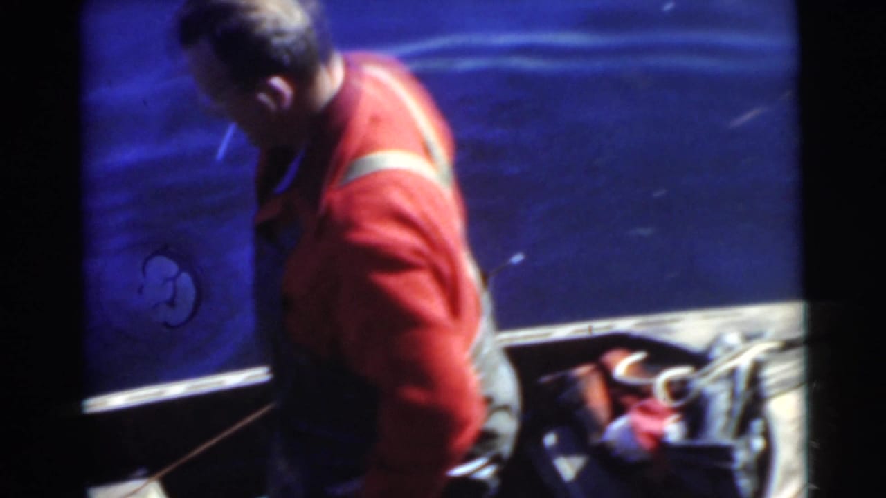 In this 1956 Miami home-movie frame, a man in a red sweater and overalls handles gear in a small open boat on blue water.