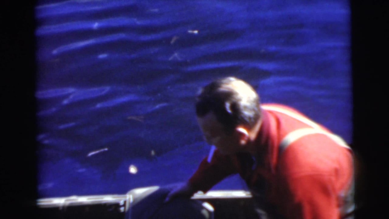 Man in a red sweater bends over the side of a small boat on blue water.