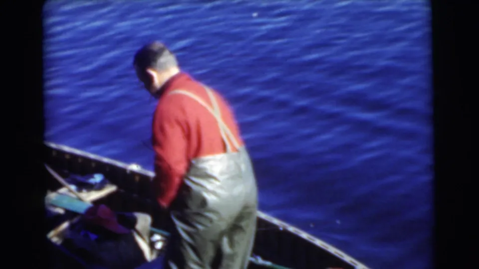 A man in overalls and a red sweater stands with his back to the camera in a small boat on Miami water in 1956.
