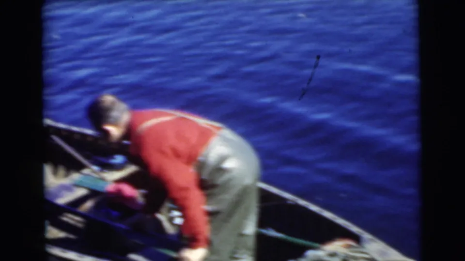A man in a red sweater and overalls bends inside a small open boat during the 1956 Miami fishing footage.