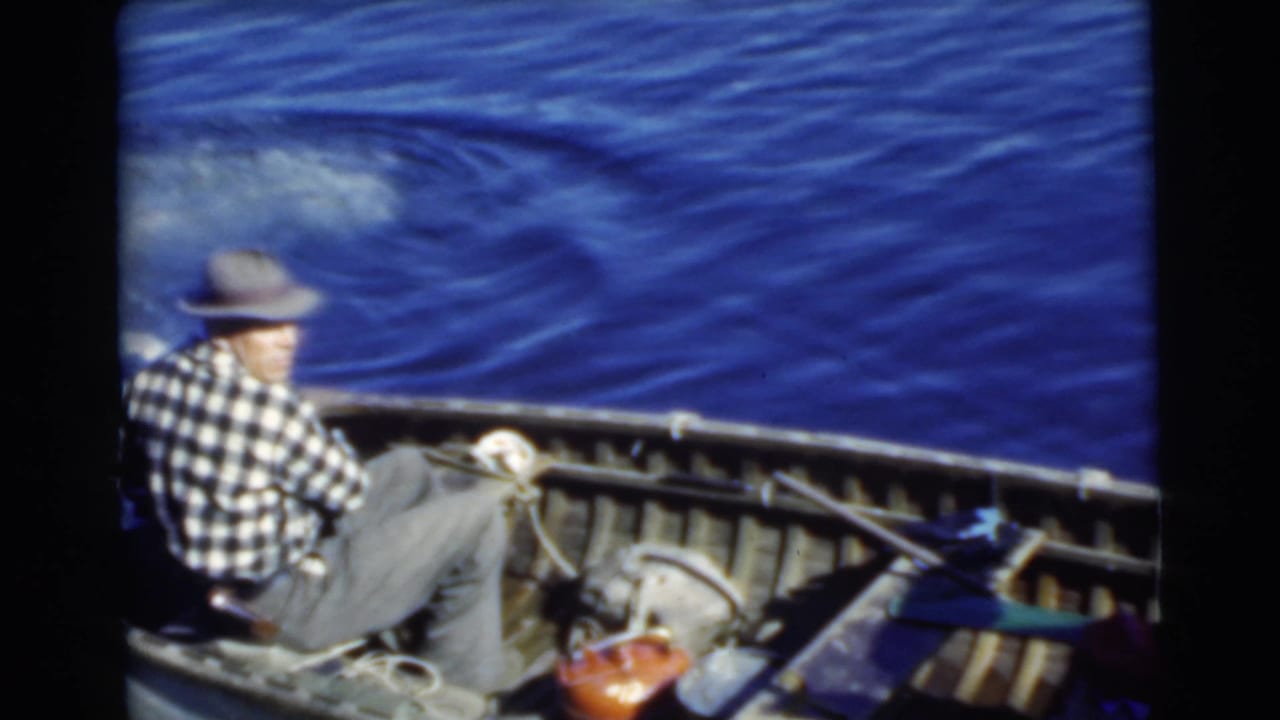 Man in fedora and checkered shirt steers a small wooden boat on blue water.