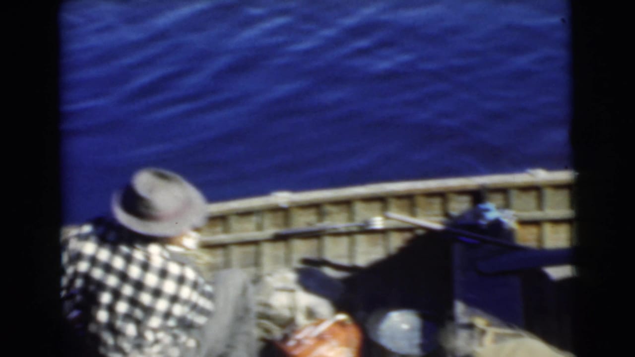 Man in fedora sits low in a wooden boat beside oars and gear on blue water.