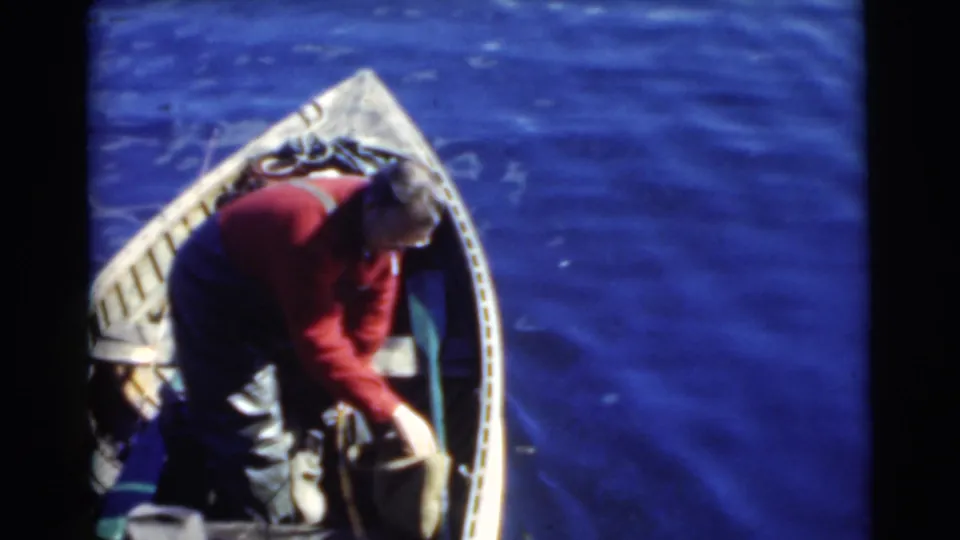 1956 Miami frame showing a man in a red shirt bent over inside a small wooden boat, possibly handling gear near the side.
