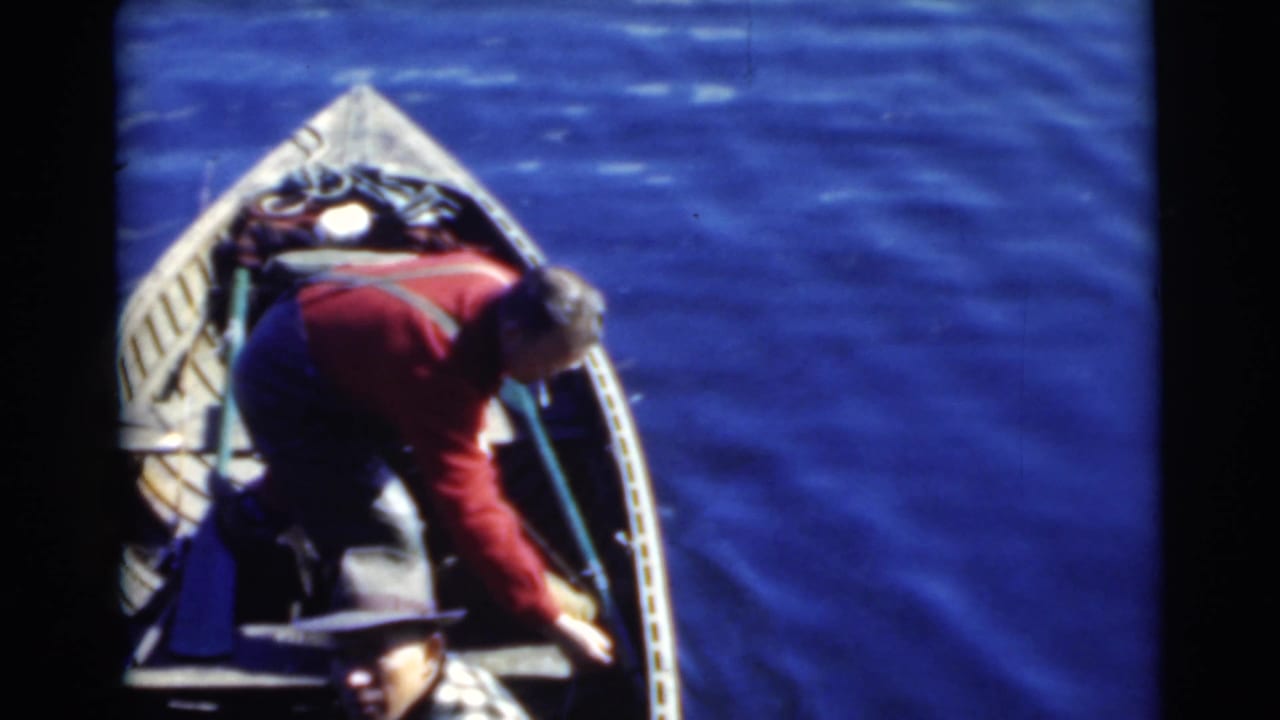 1956 Miami home-movie frame of two adult men in a small motorboat, one in a red shirt leaning toward the bow while the seated man is partly visible.