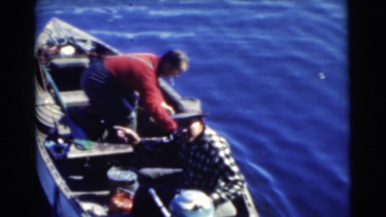 Man in red shirt leans toward an older seated man in a motorboat on calm water.