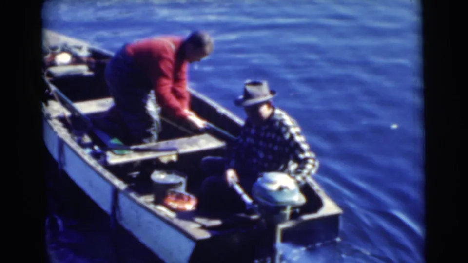 1956 Miami home-movie view of two adult men in a small motorboat, the older man in a fedora seated beside the outboard motor.