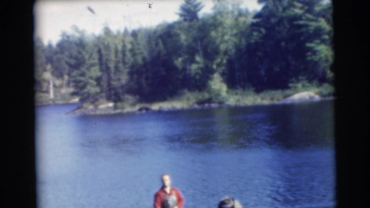 Small boat partly visible on blue lake with a man in a red shirt near the lower edge and trees across the shoreline.