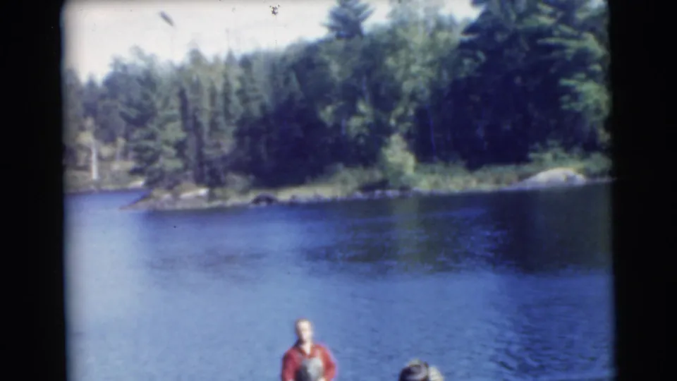 1956 8mm frame of a man in a red shirt partly visible in a small boat on blue water near a wooded shoreline.