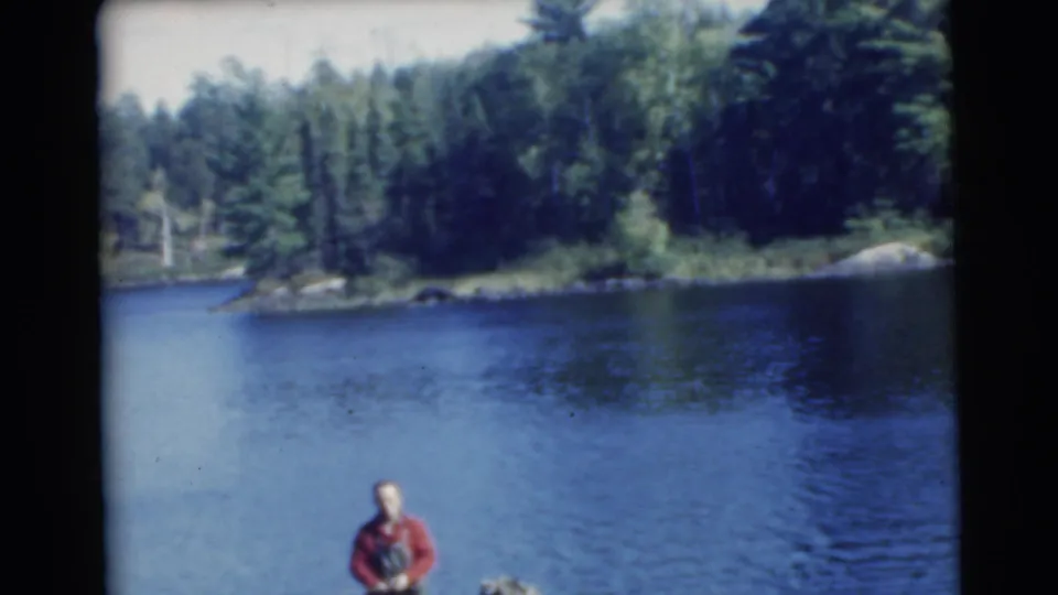 1956 8mm frame of a small fishing boat on a blue lake, with one man standing and another partly visible against a wooded shore.