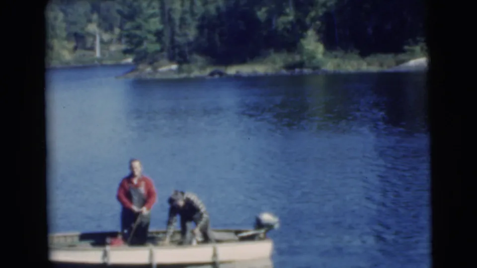 1956 8mm frame showing two adult men fishing from a small boat on blue water near a dense wooded shoreline.