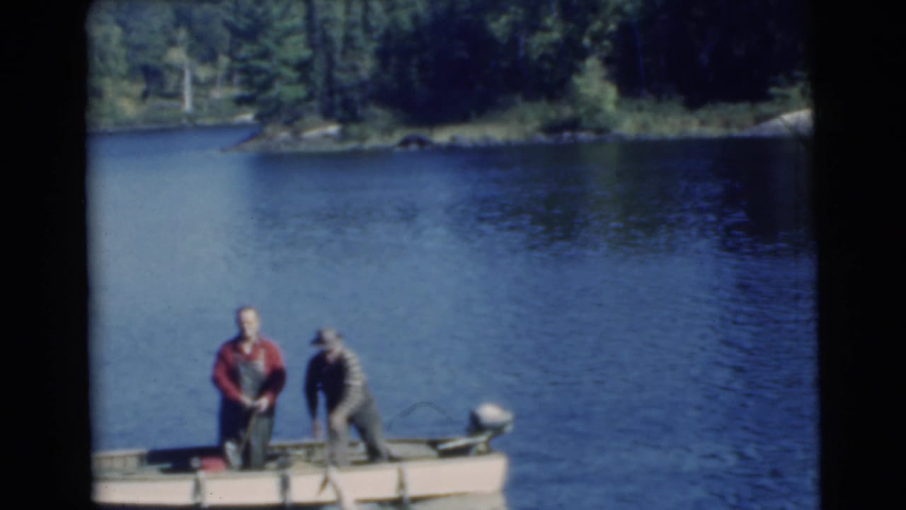 Two men stand in a small motorboat on a blue lake near a wooded shoreline.