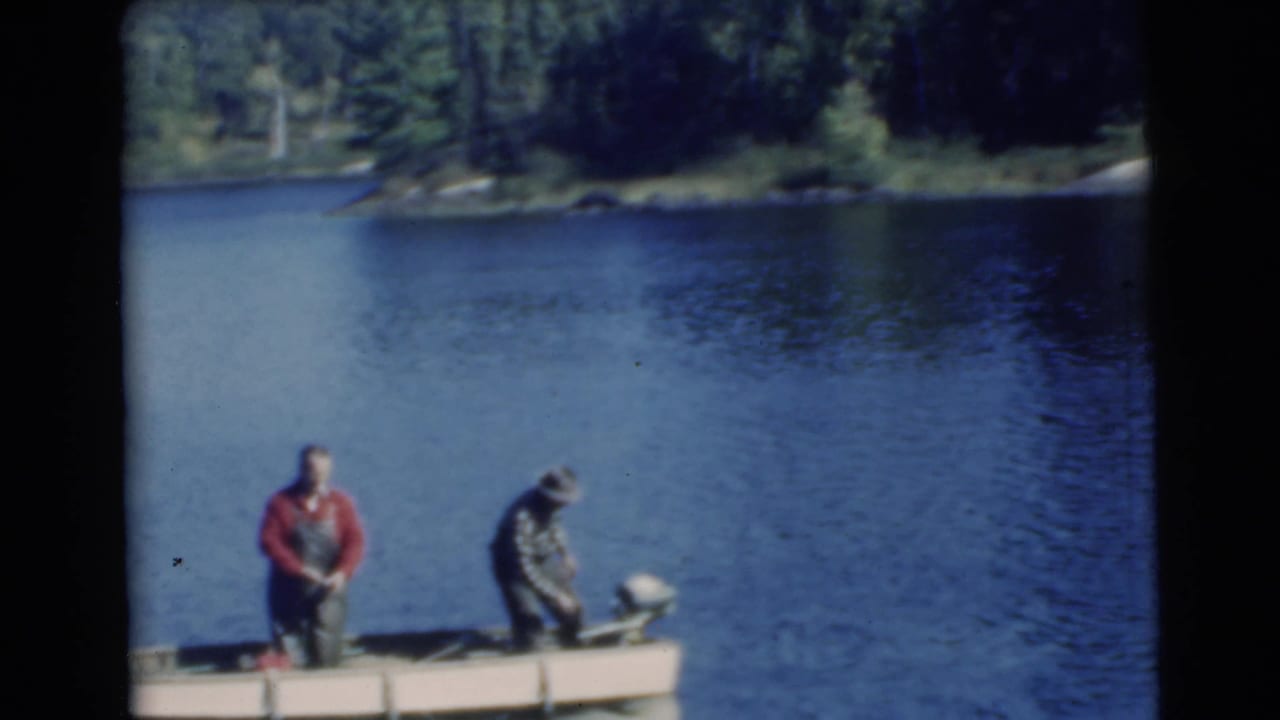 A man in red stands while another man bends near the motor at the stern of a small boat.