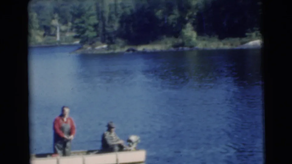 A small 1956 Miami-area motorboat sits on blue lake water as one man stands and another sits near the stern.