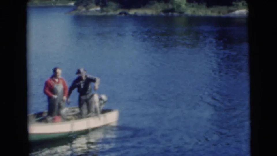 Two adult men are aboard a small motorboat on a 1956 Miami-area lake, with rippled water and forested shoreline behind them.