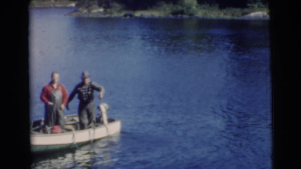 Two men stand in a small boat on blue lake water beside a large pale fish.