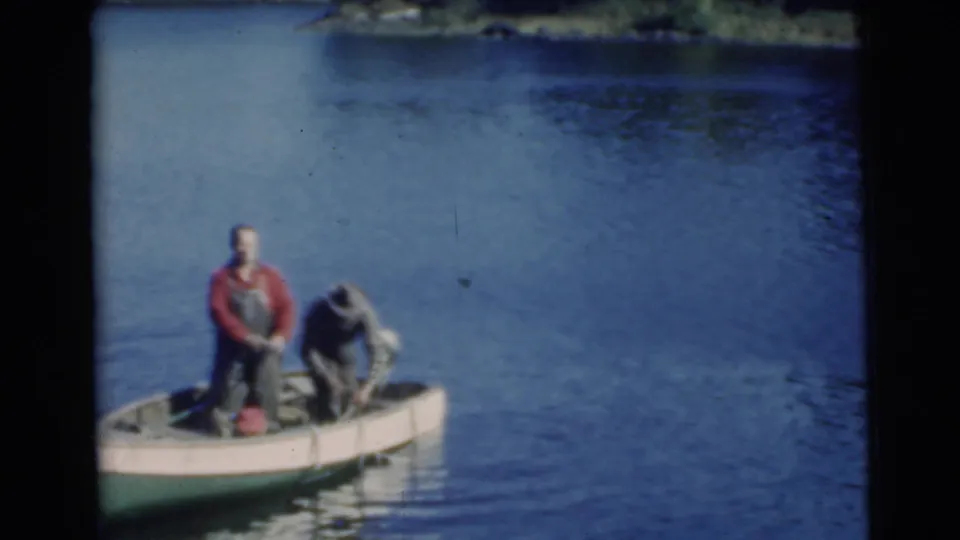 1956 Miami home-movie still with one man bending in a small boat and another standing as they handle fishing gear or a catch.