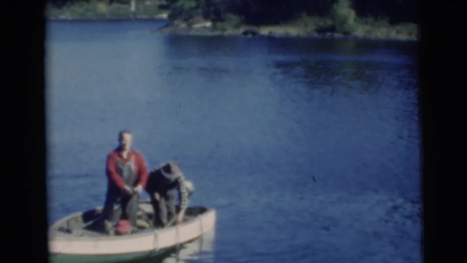 1956 Miami frame showing two adult men in a small open boat on calm lake water, one standing and one bent over near the catch area.