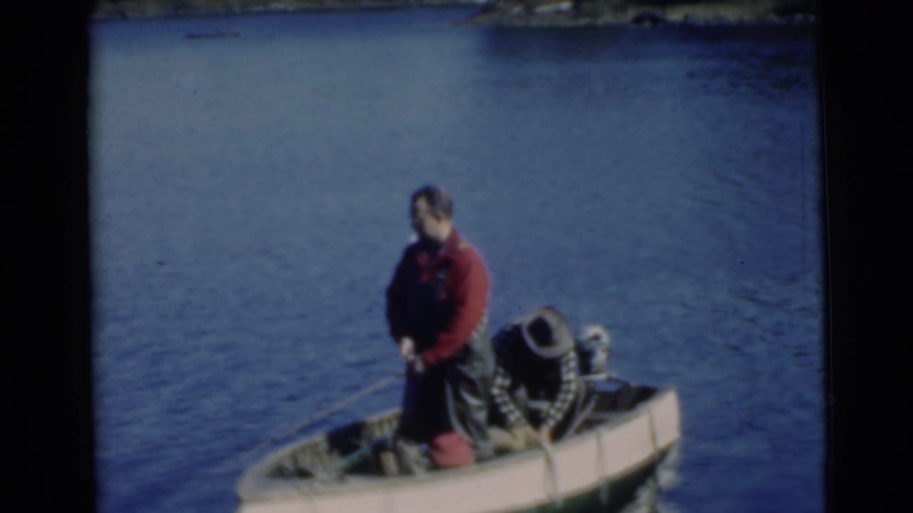 Man in red shirt and overalls standing in a small boat on a lake.