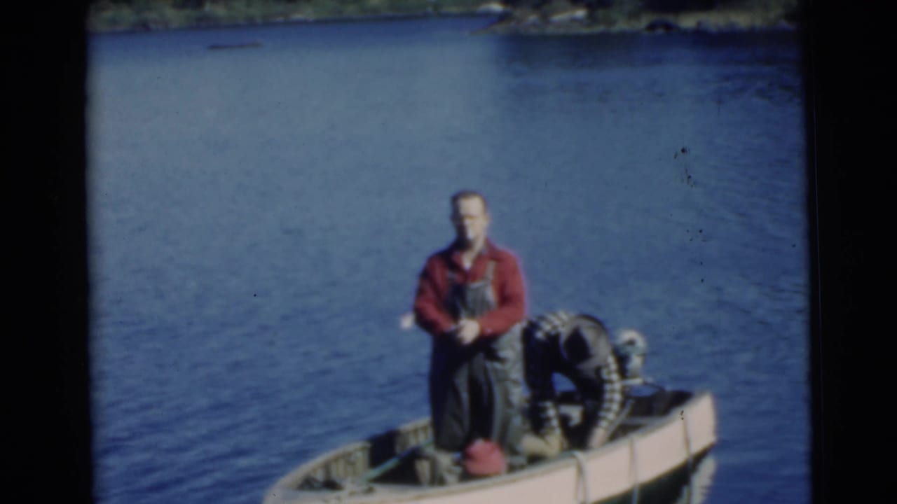 1956 Miami frame with a man in red shirt and overalls standing beside an outboard motor in a small boat on a lake.