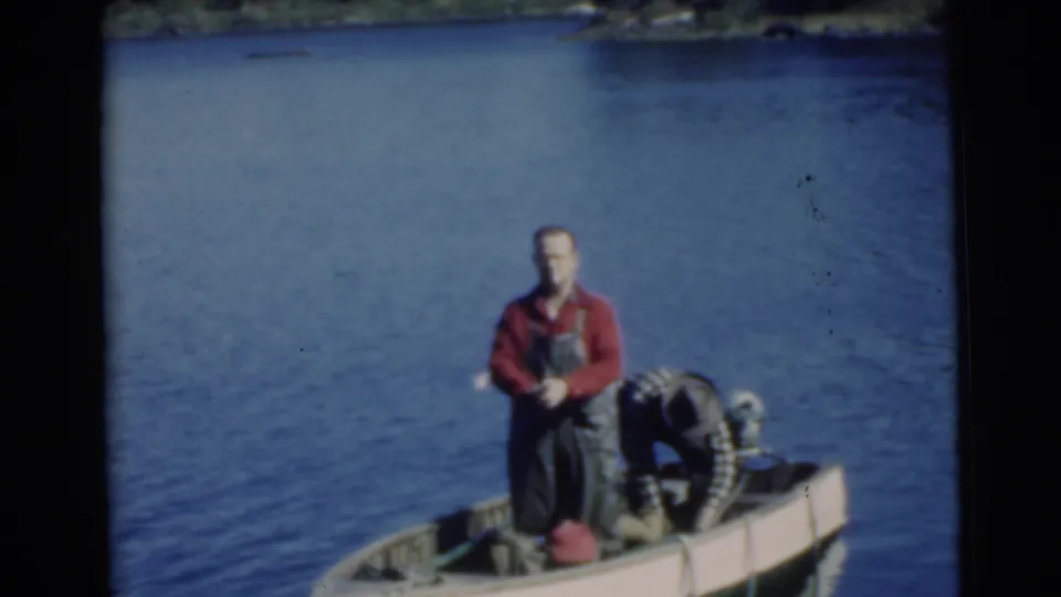 1956 Miami frame with a man in red shirt and overalls standing beside an outboard motor in a small boat on a lake.