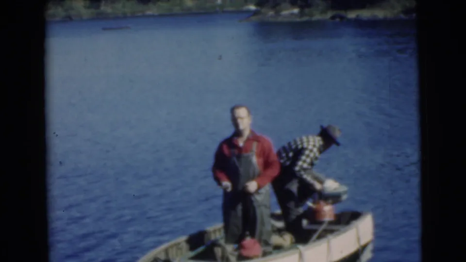 1956 Miami home-movie frame showing two adult men in a small boat, with one bending near the stern and motor equipment.