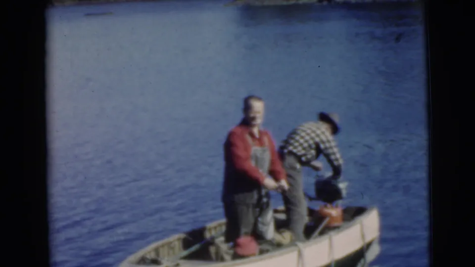 1956 Miami frame of two men aboard a small boat on calm water, one standing while the other tends equipment near the outboard motor.