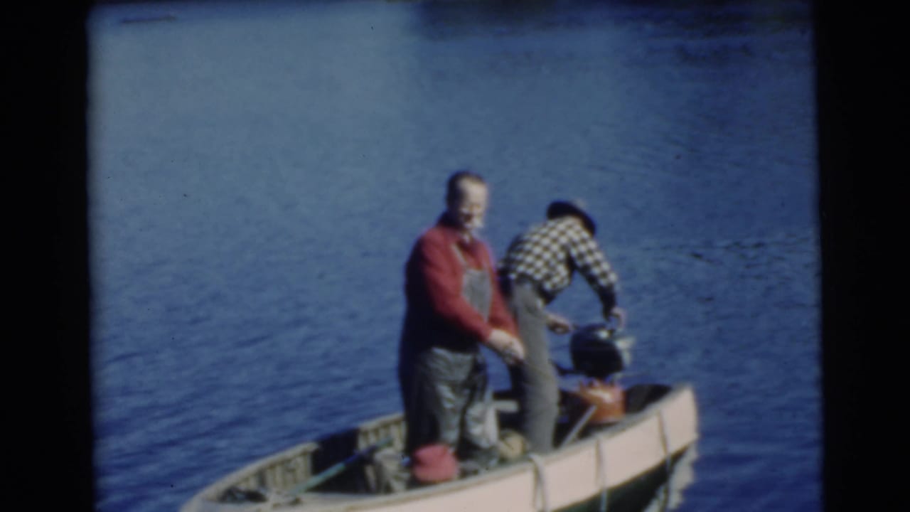Two adult men stand in a small boat on a lake, one holding a fishing rod while the other tends to gear.
