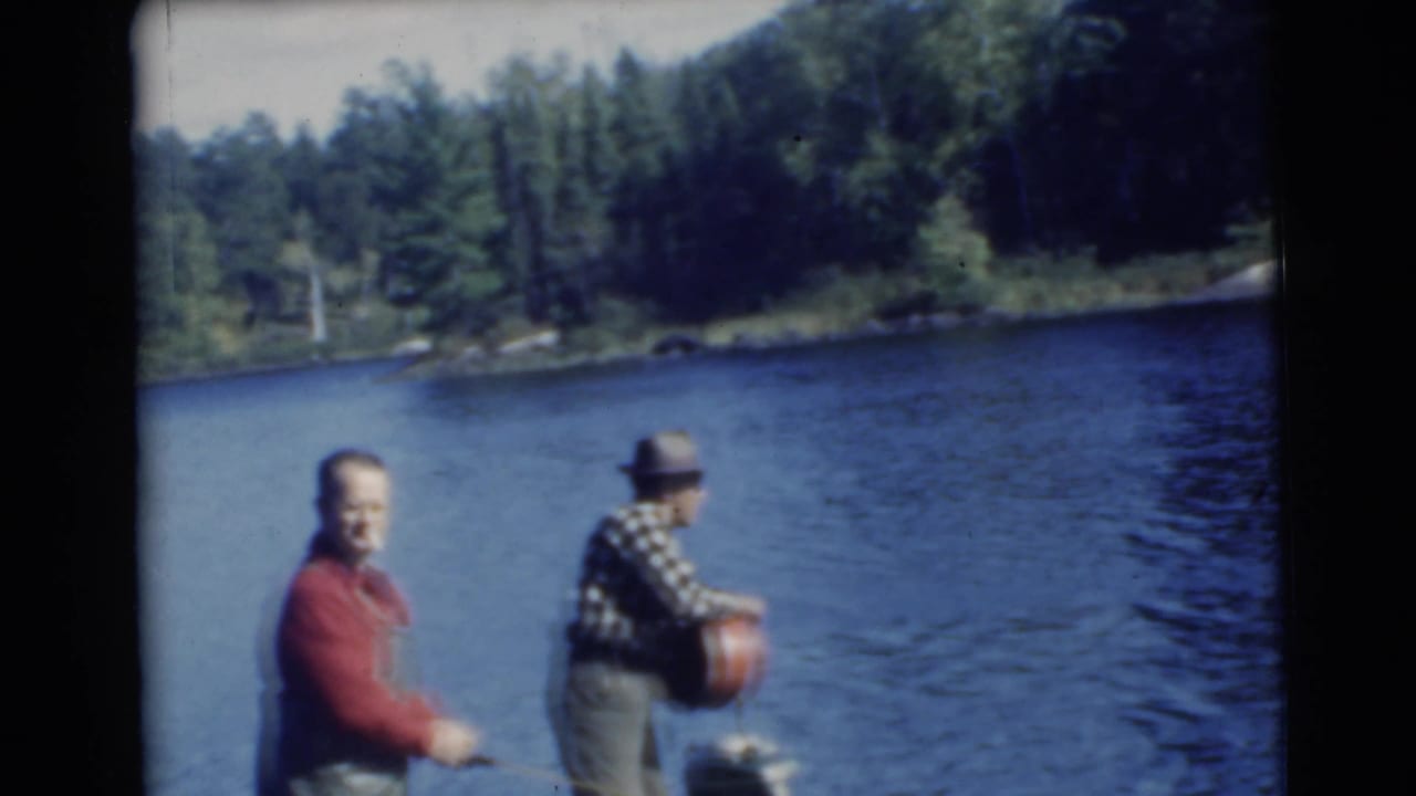 A man in a red shirt faces the camera while another man in a hat stands near boat gear on the lake.