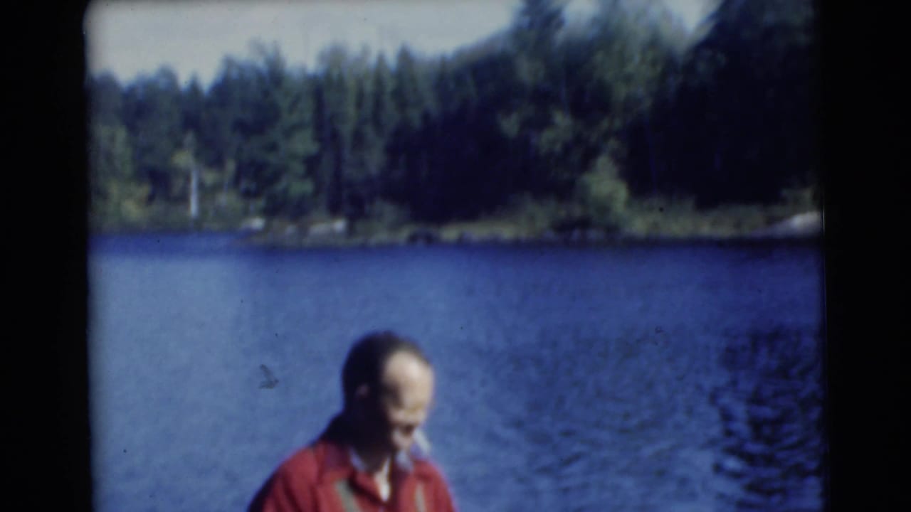 Adult man in a red shirt stands near a blue lake with wooded shoreline behind him.