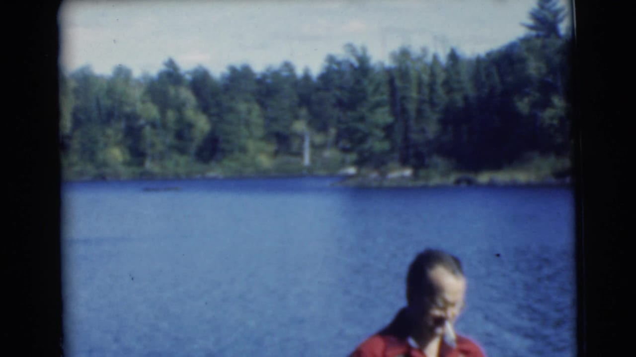 Man in a red shirt appears at the edge of the frame beside a lake and wooded shore.
