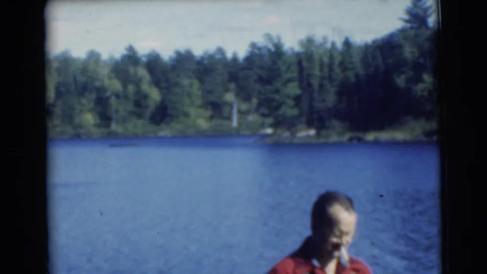 Adult man in red is partially framed beside blue lake water and a tree-lined shoreline in 1956 home-movie footage.