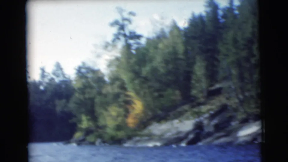 1956 8mm lakeside view showing rippled blue water, sloped rock, and dense green trees along the shore.