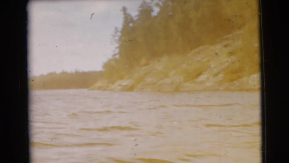 1956 8mm home-movie view across rippling water toward a rocky, tree-covered shoreline with faded warm color.