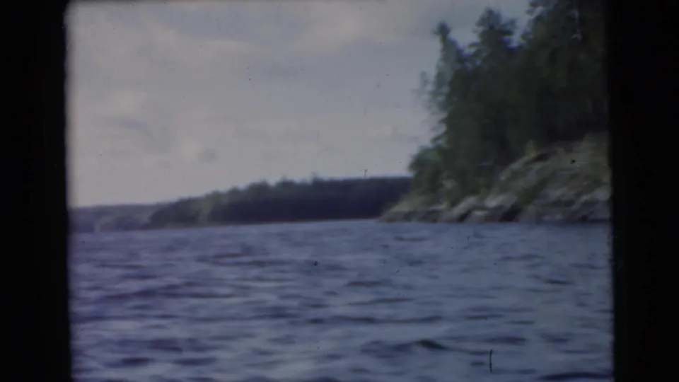 1956 8mm frame with blue-toned rippling water, distant wooded shore, rocky right bank, and dark film edges.