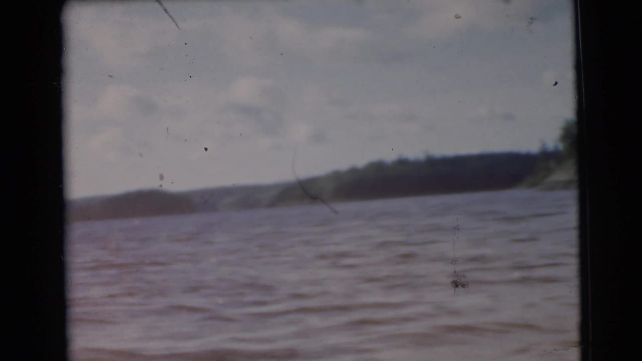 1956 Miami home-movie frame shows rippling lake water with a dark wooded shoreline and low hills in the distance.