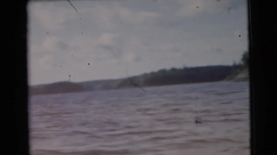 1956 Miami home-movie frame shows rippling lake water with a dark wooded shoreline and low hills in the distance.