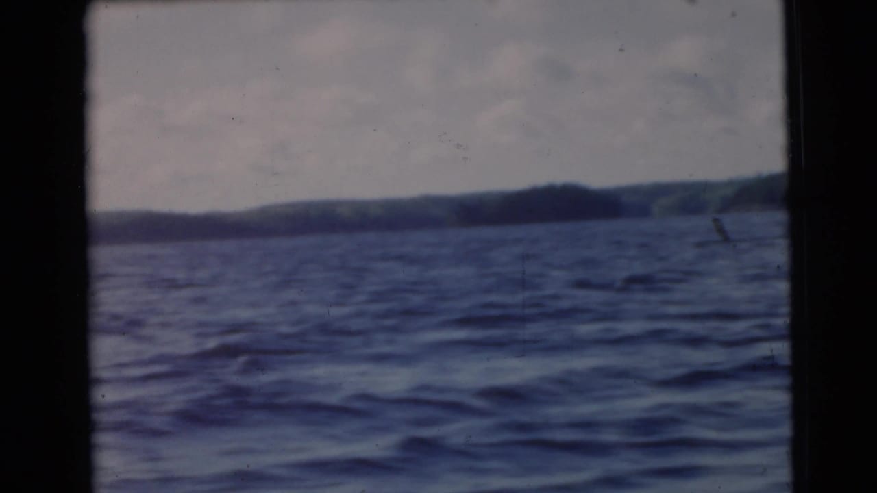 Boat-level 8mm frame from 1956 shows a broad lake surface, distant wooded hills, cloudy sky, and light film scratches.