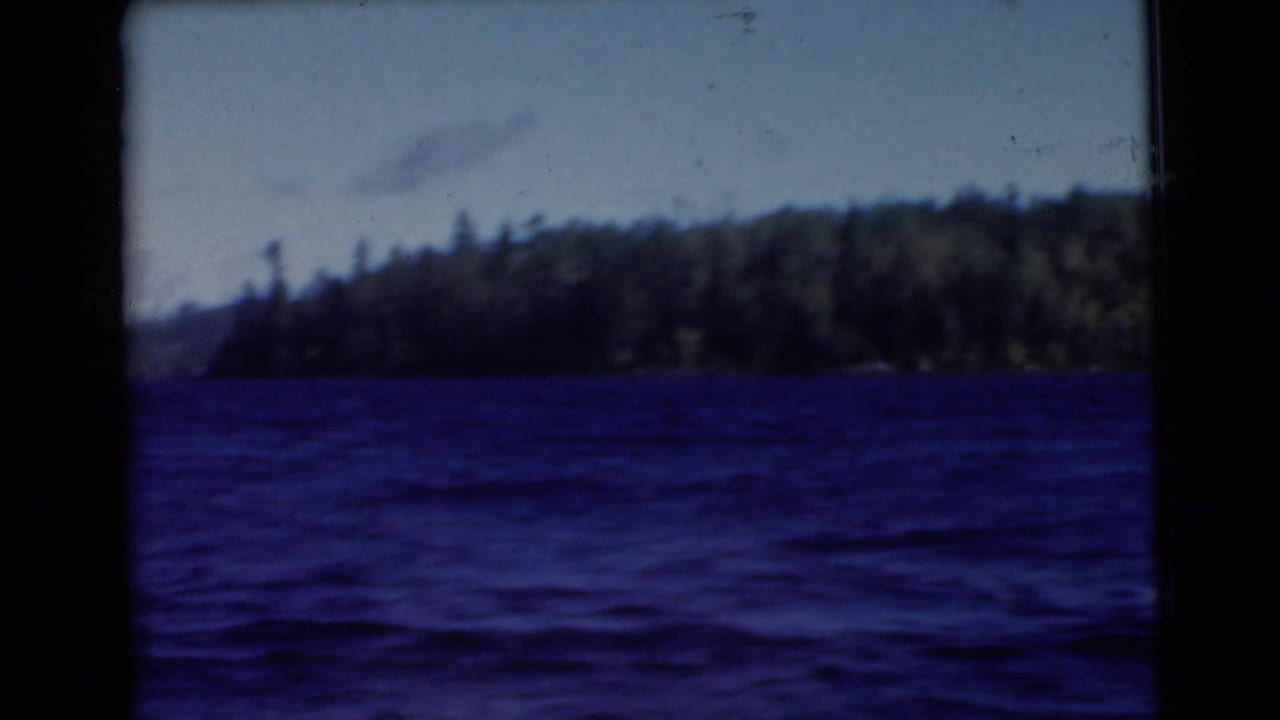 1956 8mm footage shows a blue-toned lake from a moving boat, with dense trees along the far shoreline.