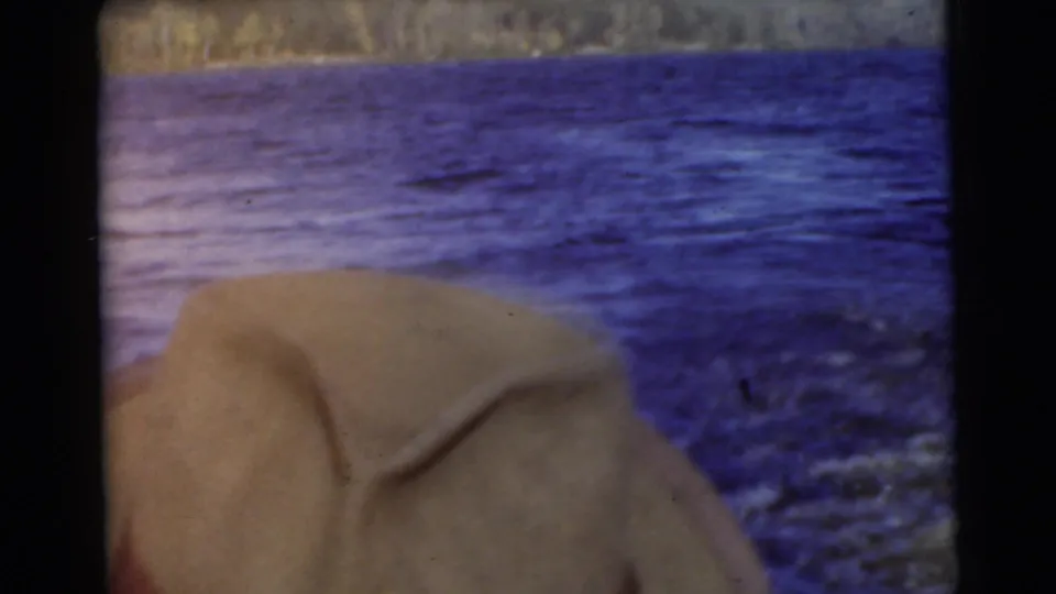 1956 Miami home-movie frame showing a light-colored hat in the foreground, agitated blue water, and a distant treeline.