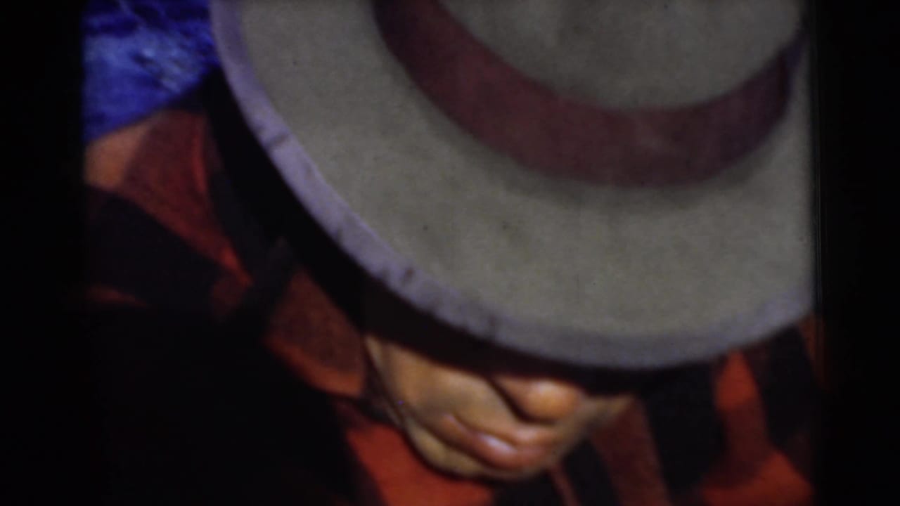 1956 Miami home-movie frame of an adult man in a light fedora and red plaid shirt, his face partly shaded as he looks down.