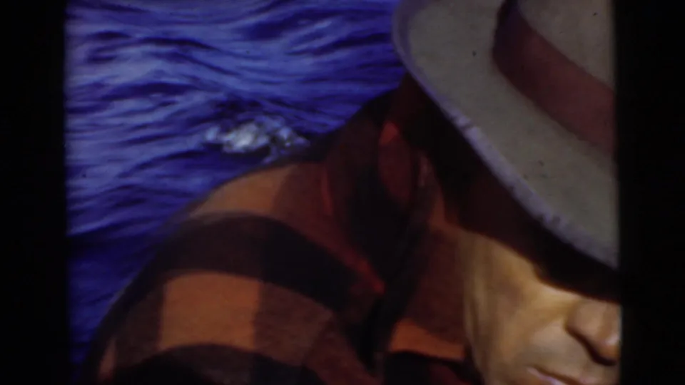 1956 Miami home-movie frame showing the man close to camera on a boat, with blue water behind him and his hat obscuring part of his face.