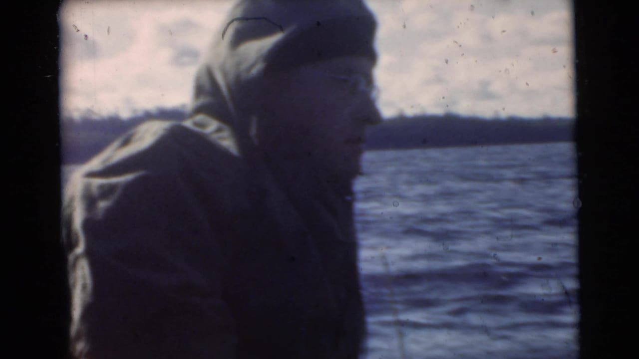 Man in a hooded jacket beside open water, seen in soft faded 8mm color.